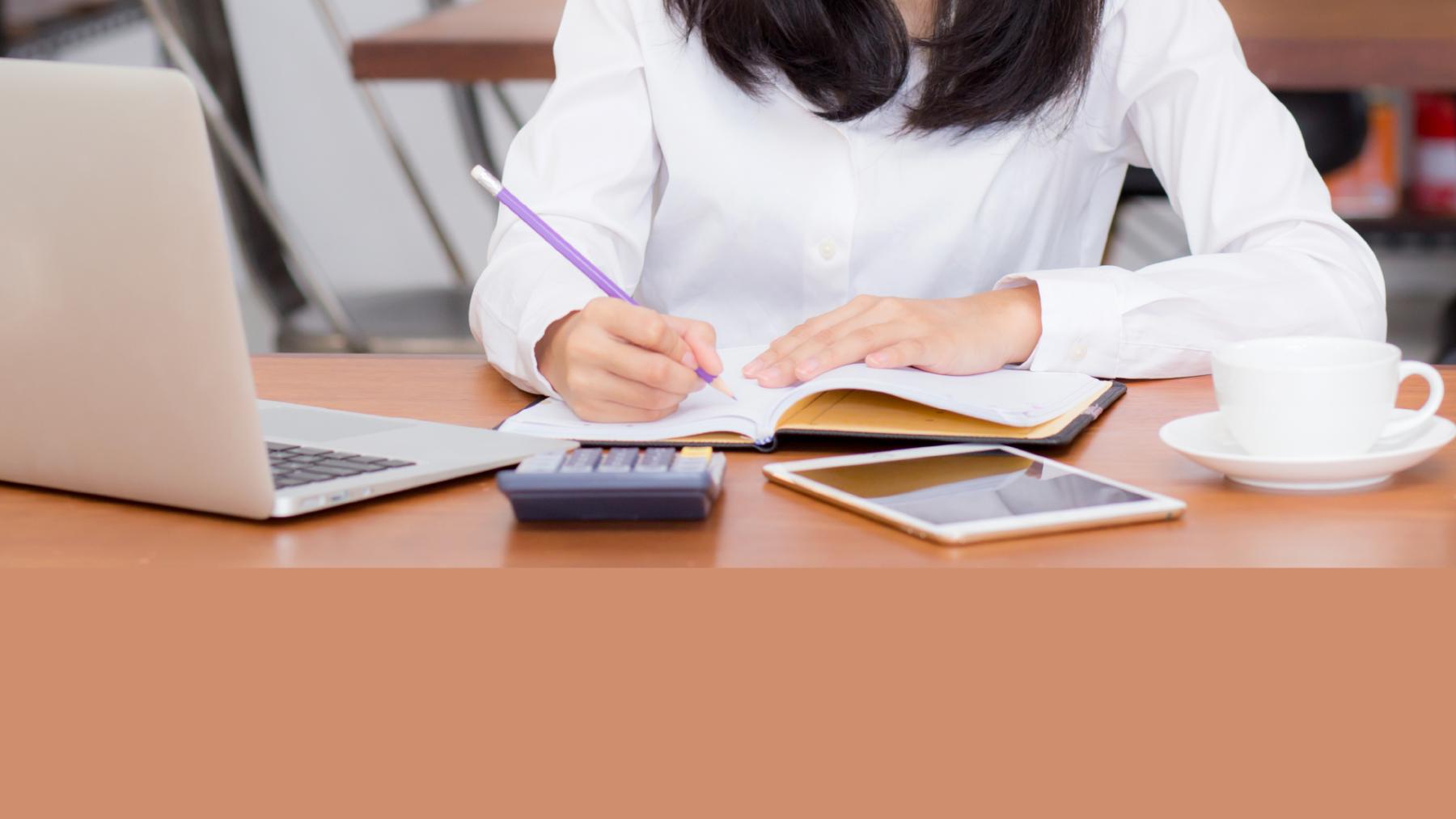 Woman writing on a desk with laptop and iPad in front of her